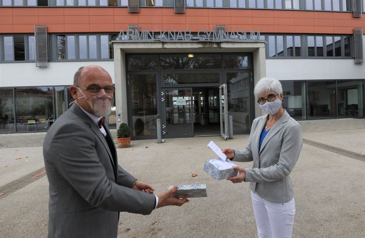 Schulreferent Jürgen Engel übergibt eine der gespendeten transparenten Coronamasken an Oberstudienrätin Anette Gerhard, die sich in der Schulpastoral am Kitzinger Armin-Knab-Gymnasium engagiert.
